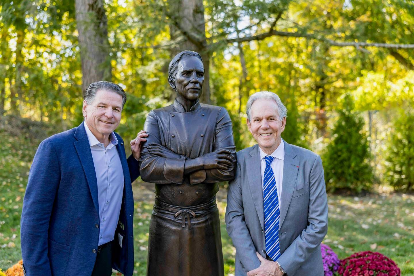 Two guests pose beside the finished bronze, which shows Keller with arms crossed in his chef's coat and tied apron.