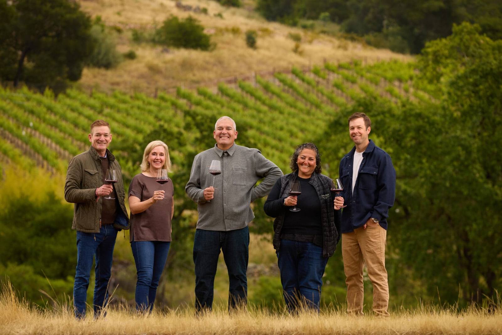 The five-member master blending team for the 2024 Collaboration stands in a Stags Leap District vineyard with glasses of the blend. The team changes every vintage, so more of the district winemakers shape the wine over time.